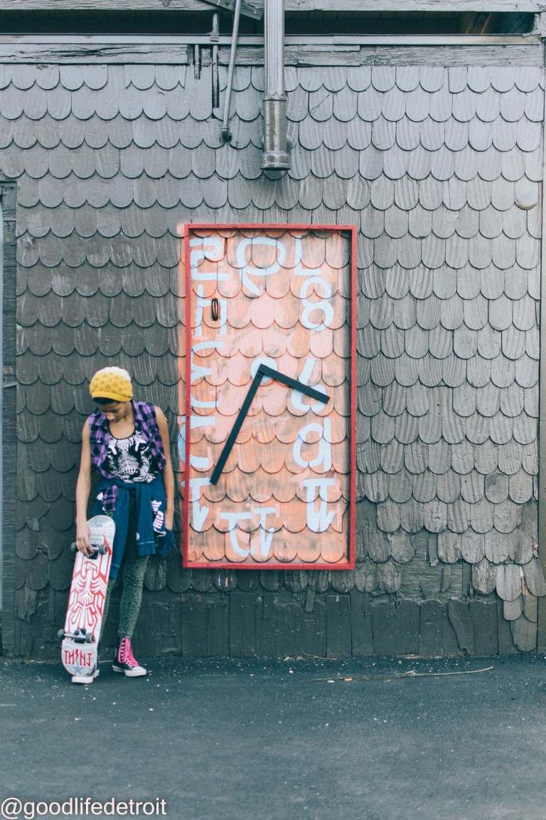 Young girl with a skateboard against a wall wearing a yellow hat and mixed patterned clothing.