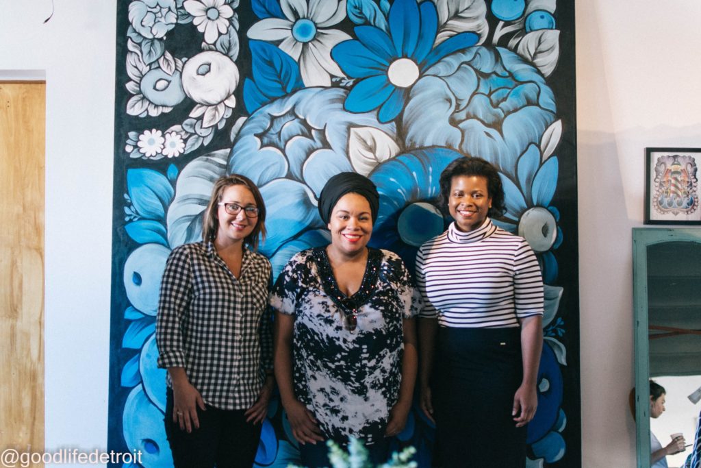 Three women stand in front of a blue floral mural that resembles the Goodwill logo.